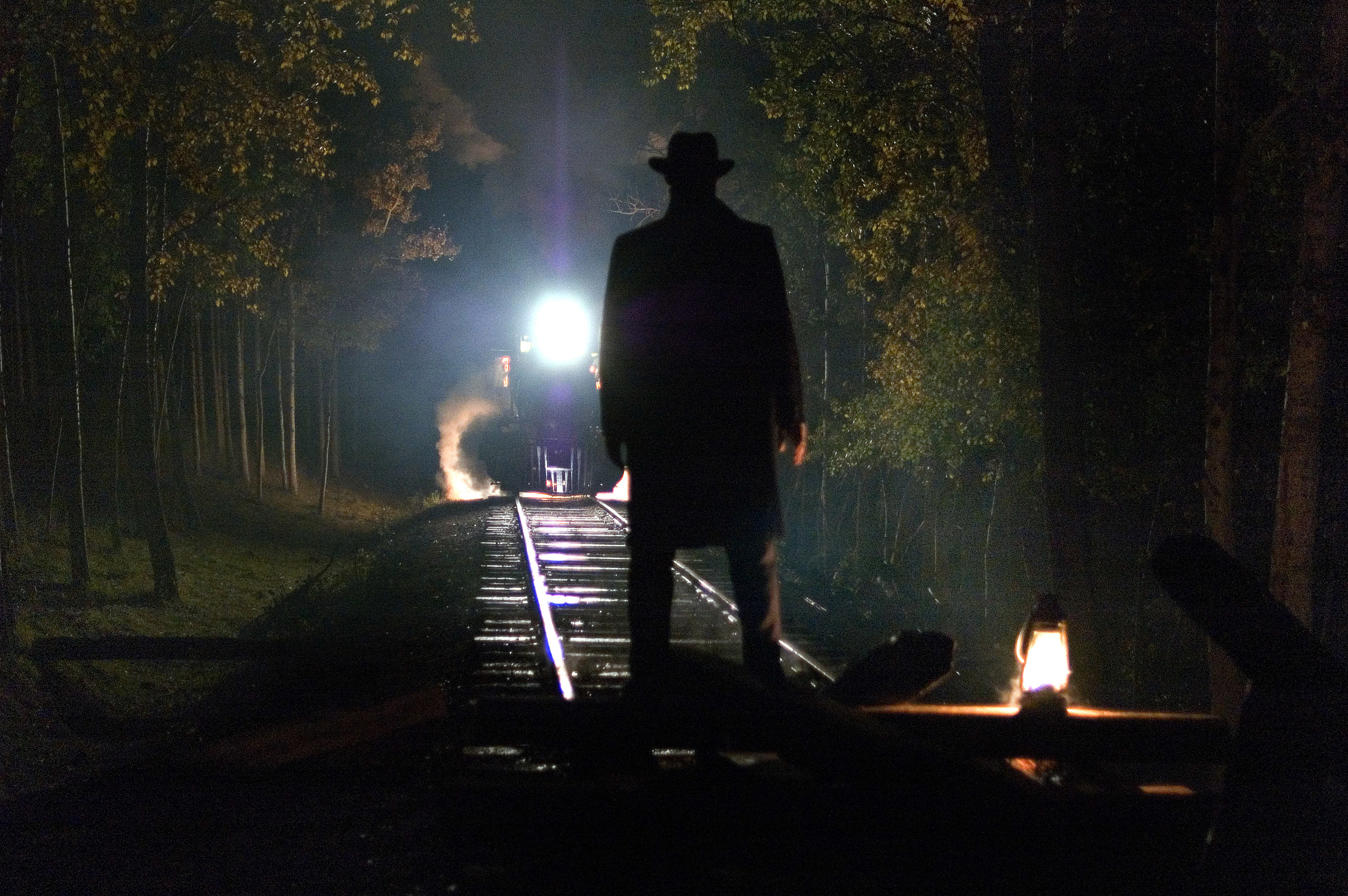 the backside of a man wearing a hat and standing in the middle of train tracks in the dark while a train moves toward him