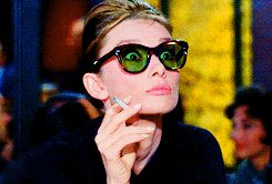 A woman with a cigarette in her fingers, with wide eyes, pulls down her sunglasses.