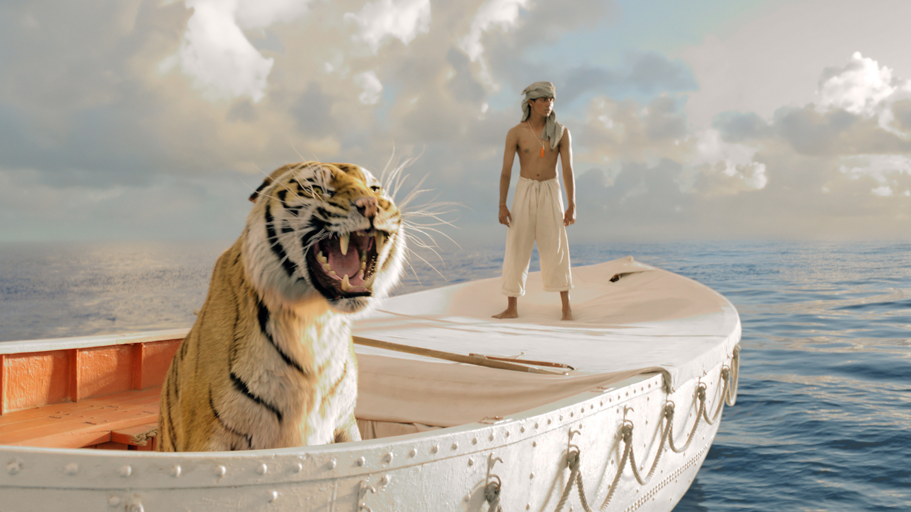 a serene shot of a tiger on a large canoe-like boat with a man standing toward the back with his shirt wrapped around his head