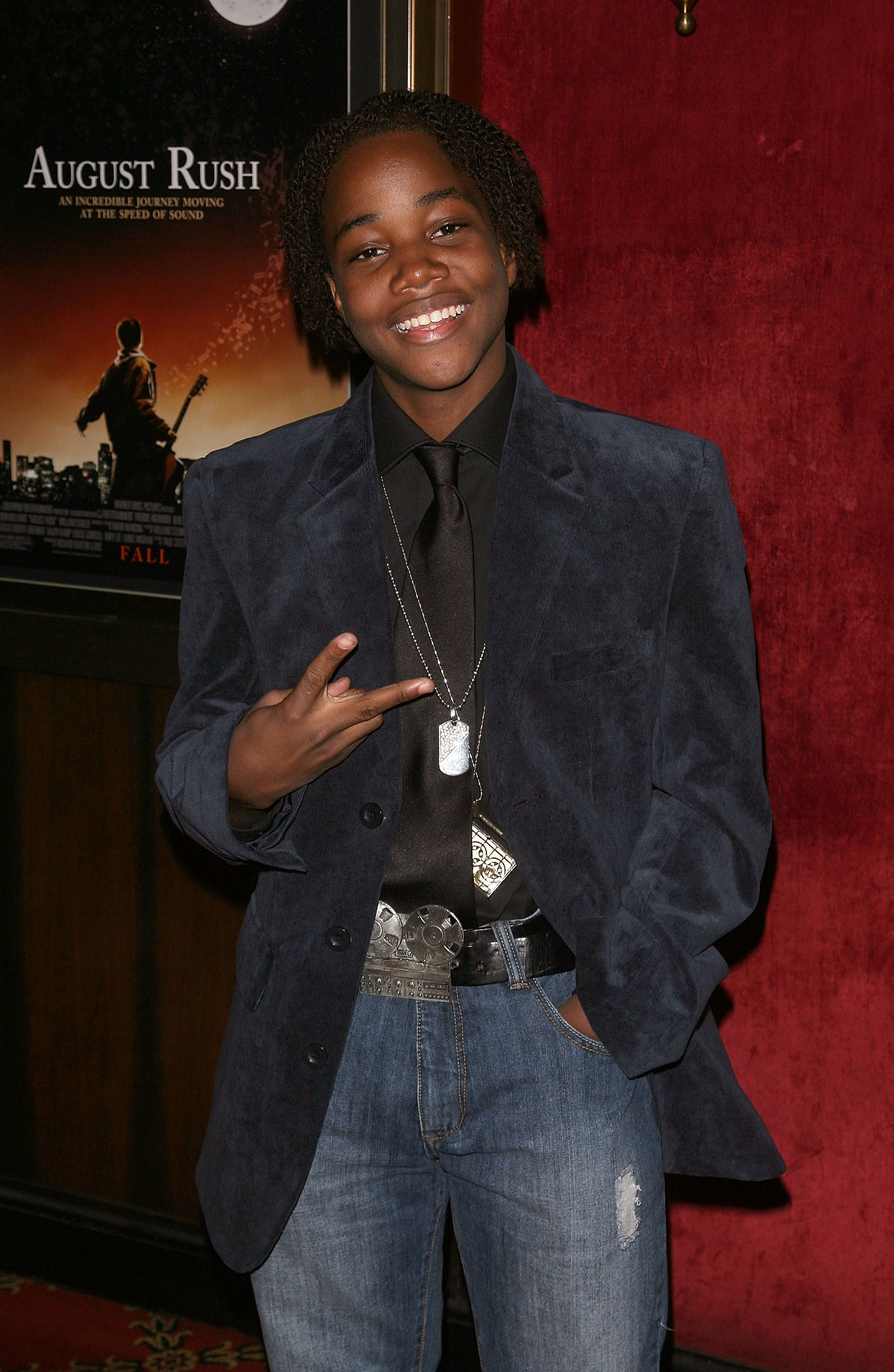 Smiling Leon giving the peace sign and wearing jeans, a tie and shirt, and a jacket