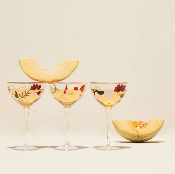 Three of the champagne coupes with liquid in them, a slice of cantaloupe balanced on top of two of them, and a cantaloupe quarter in the background