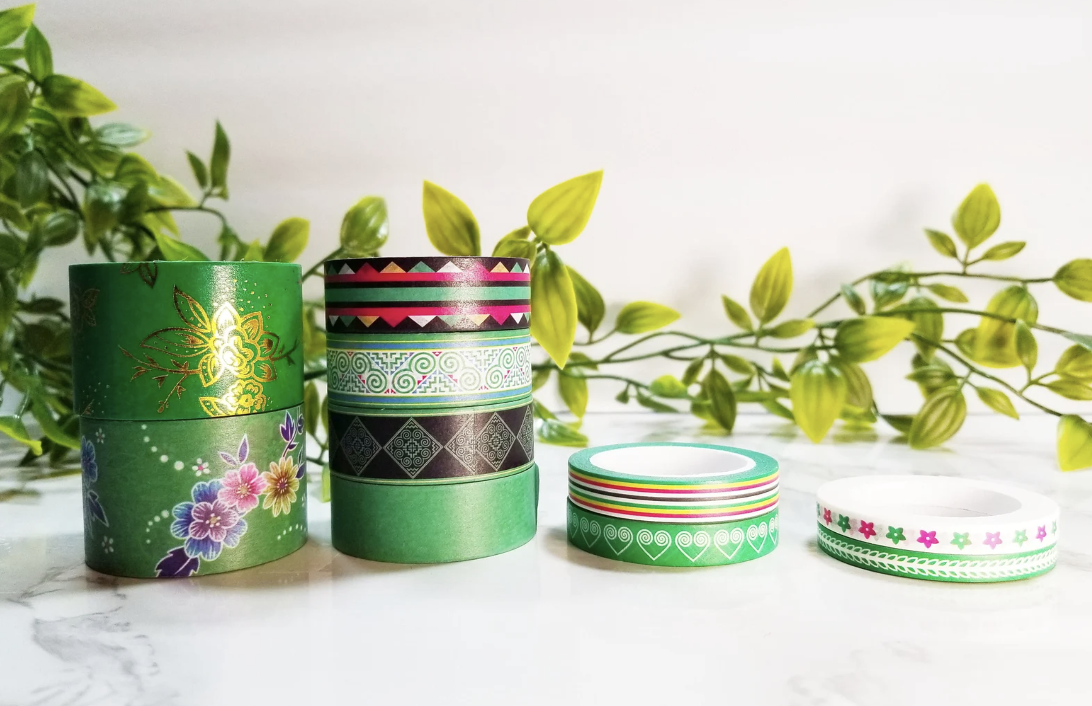 Stack of several washi tapes with floral and Hmong inspired designs in front of green leaves.