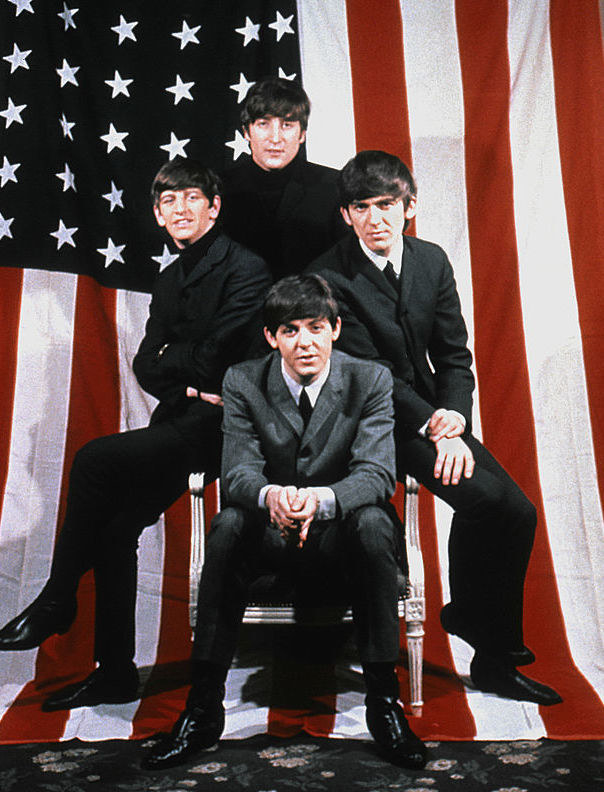 The Beatles posing for a portrait in 1964 in front of a US flag