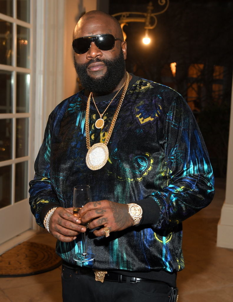 Rick Ross wearing sunglasses, standing and holding a glass of champagne