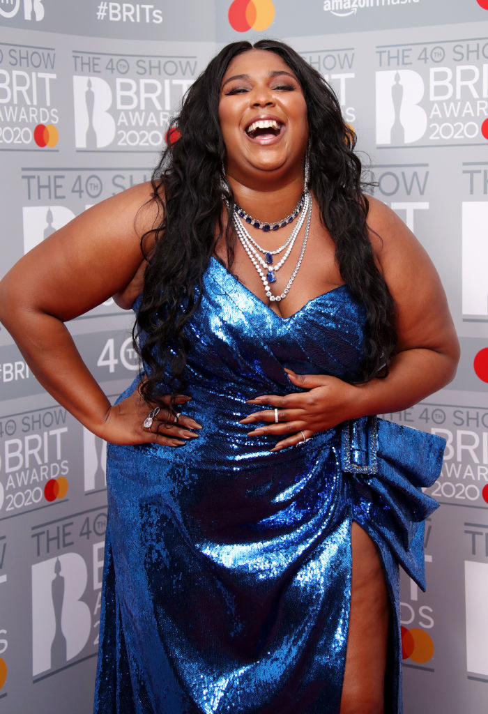 Lizzo smiling, with her hands on her waist, wearing a shiny, high-slit gown