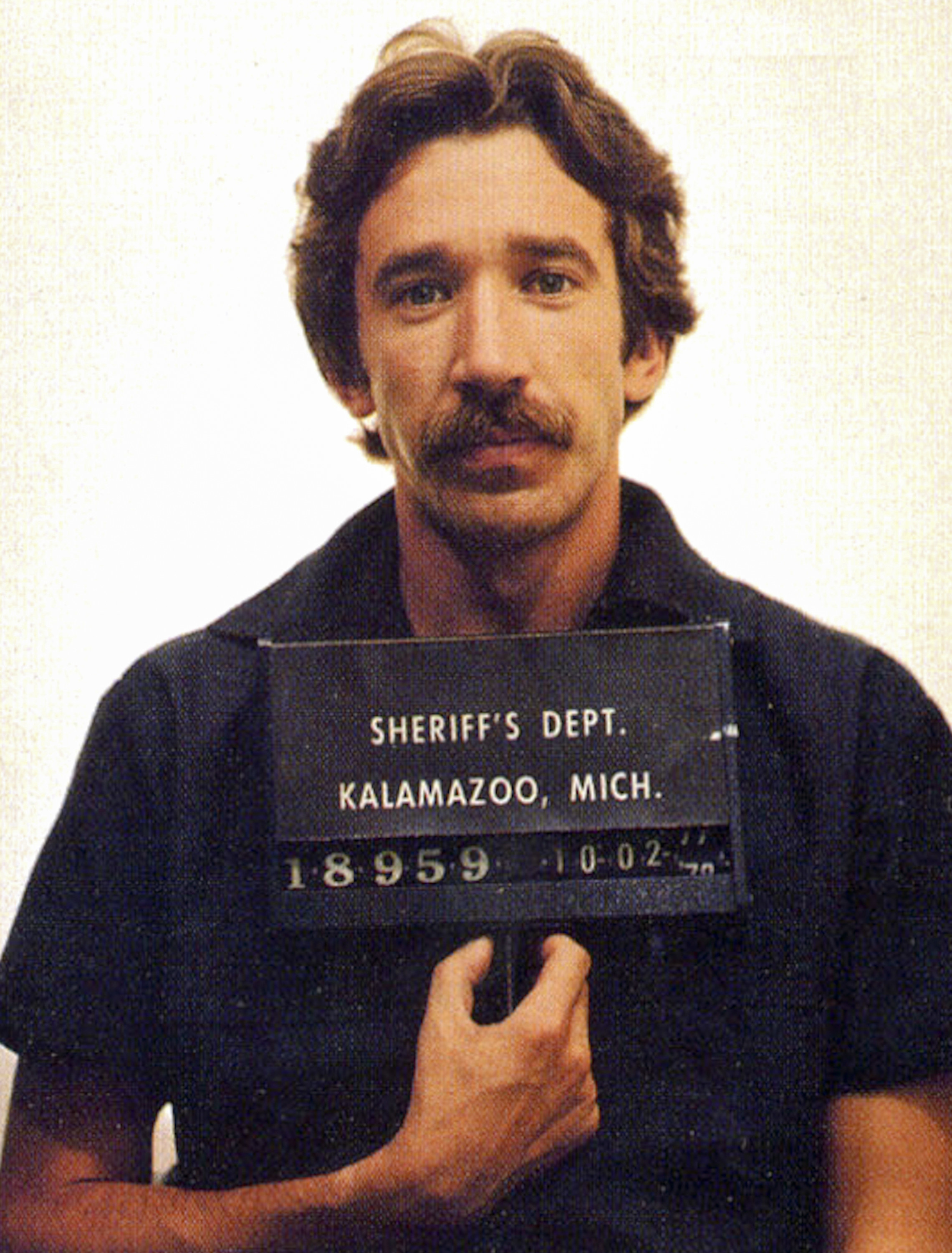 A young, mustachioed Tim Allen holding a ID placard in a mugshot
