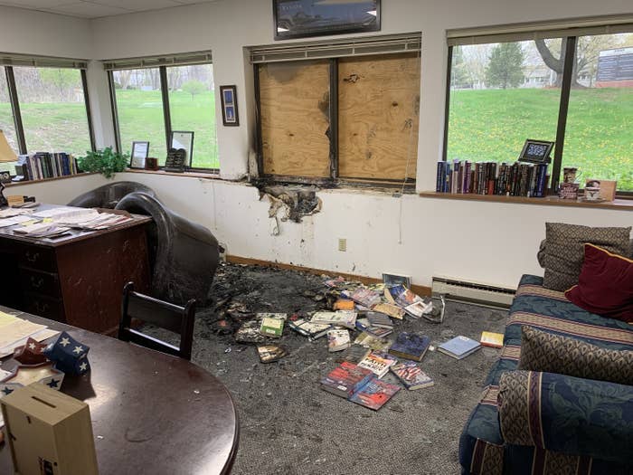 An interior of the office shows charred floors and walls, burned books, and a broken window covered up with wood