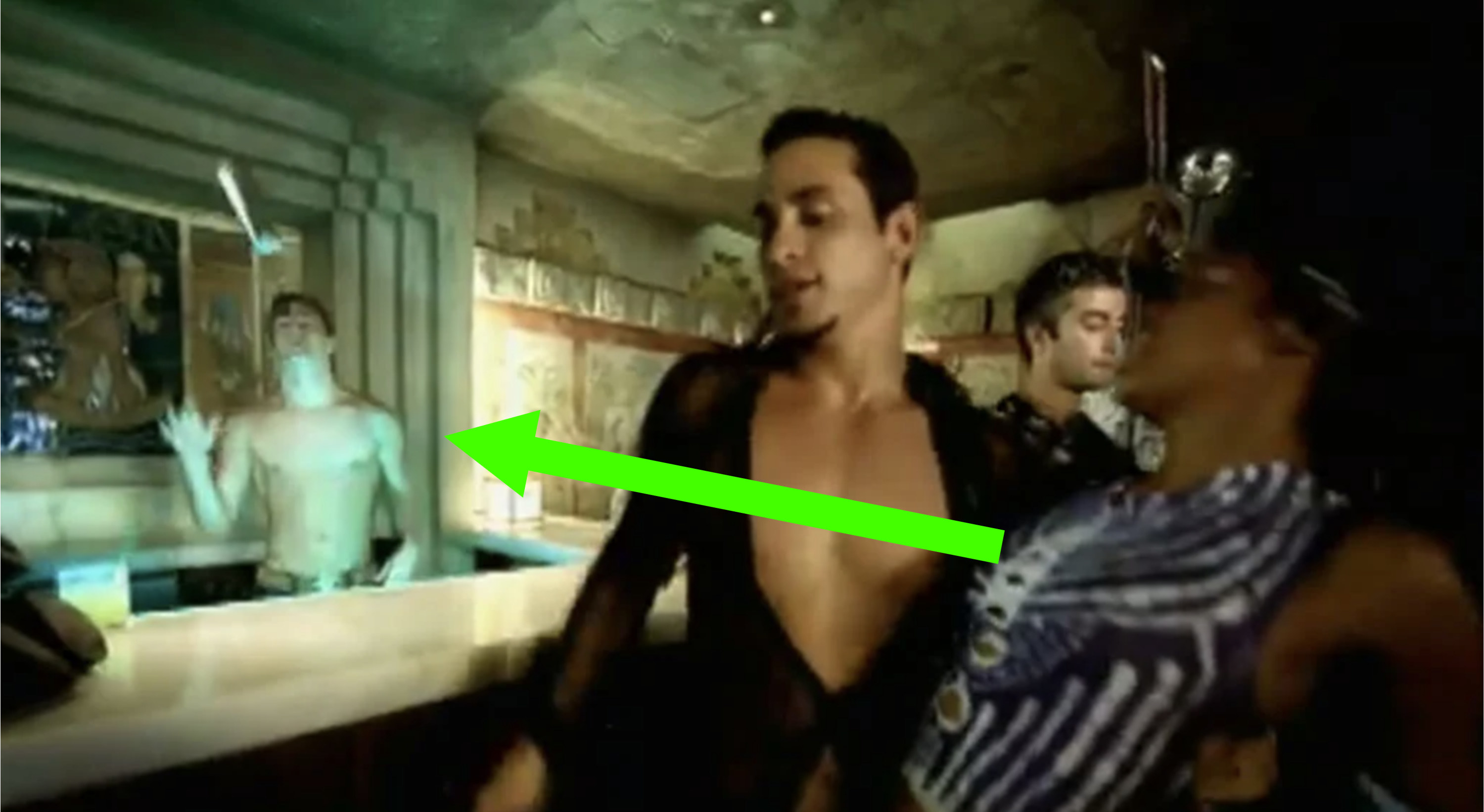 A still from a music video, set in a bar, there&#x27;s a shirtless bartender throwing a cocktail shaker and an arrow pointing at him