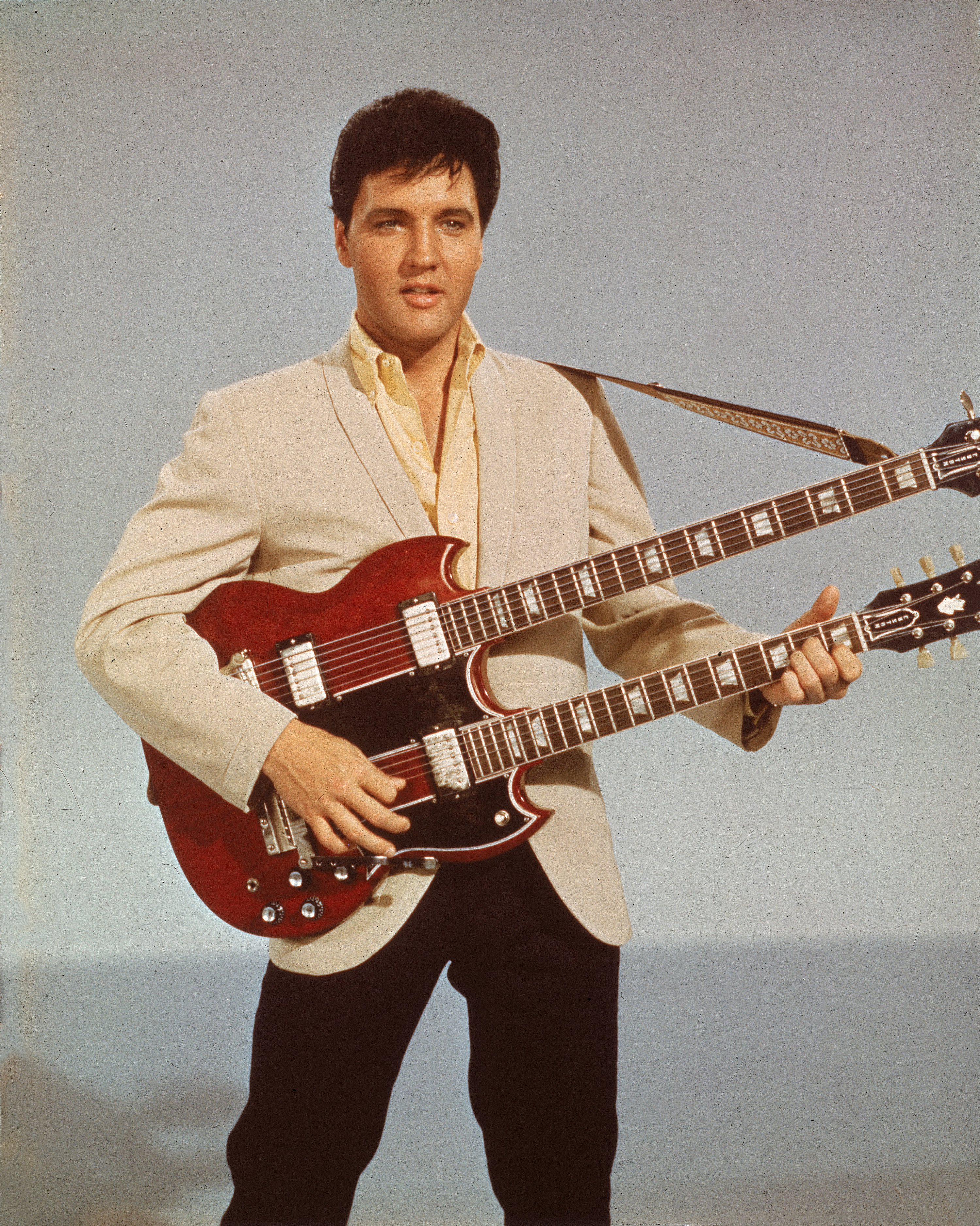 Elvis Presley wears a white jacket and smiles while holding a double-necked guitar