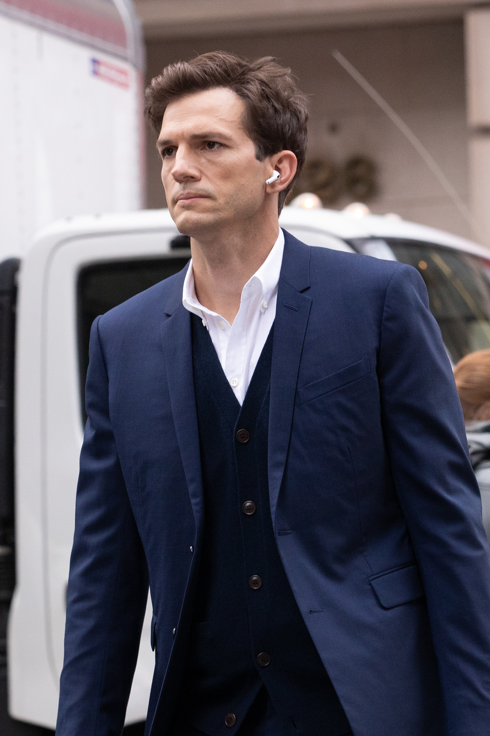 Ashton Kutcher walks down a city street in a blue suit, with Airpods in
