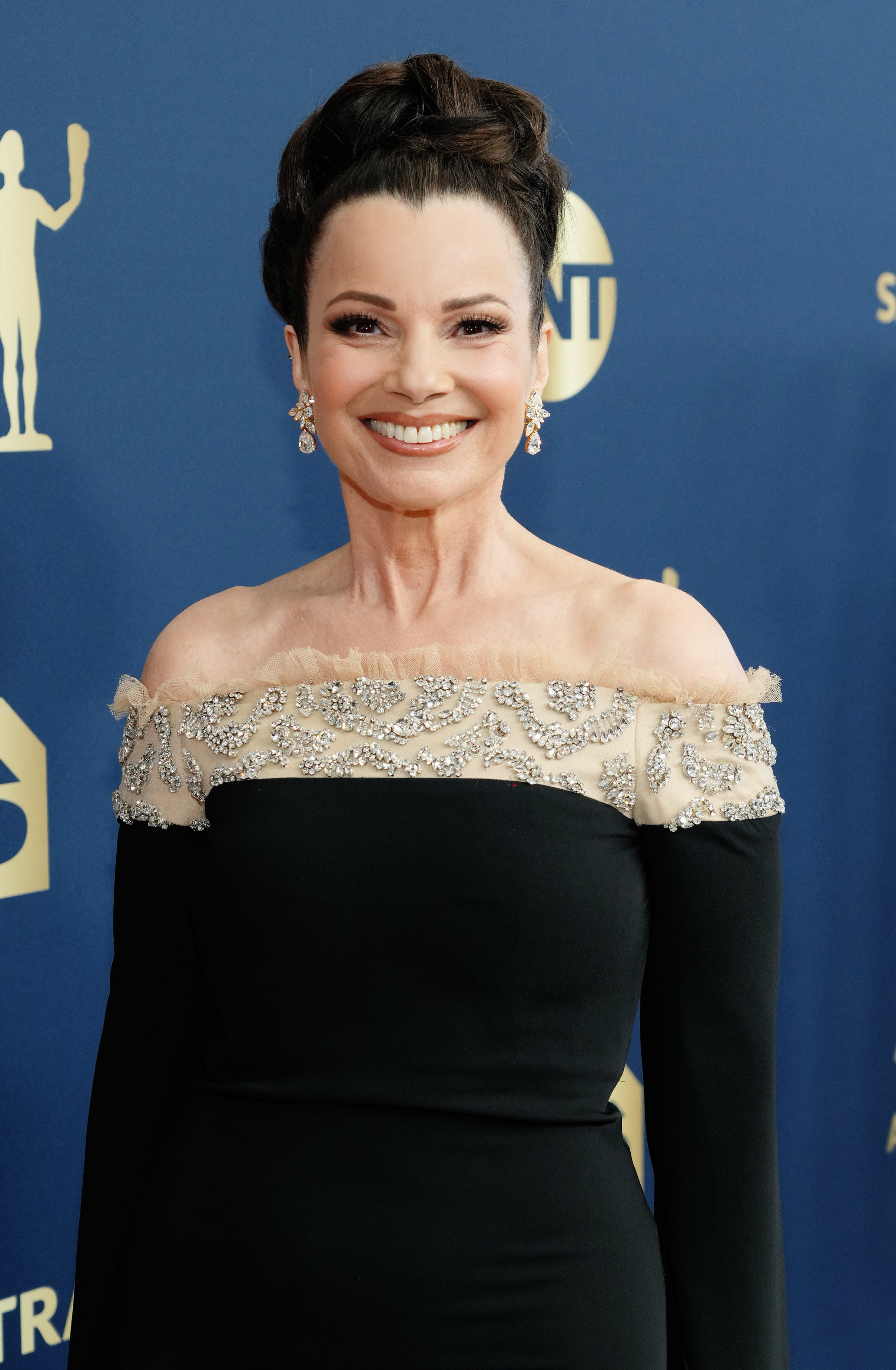 Fran Drescher smiles while on the red carpet wearing a black and cream off the shoulder dress