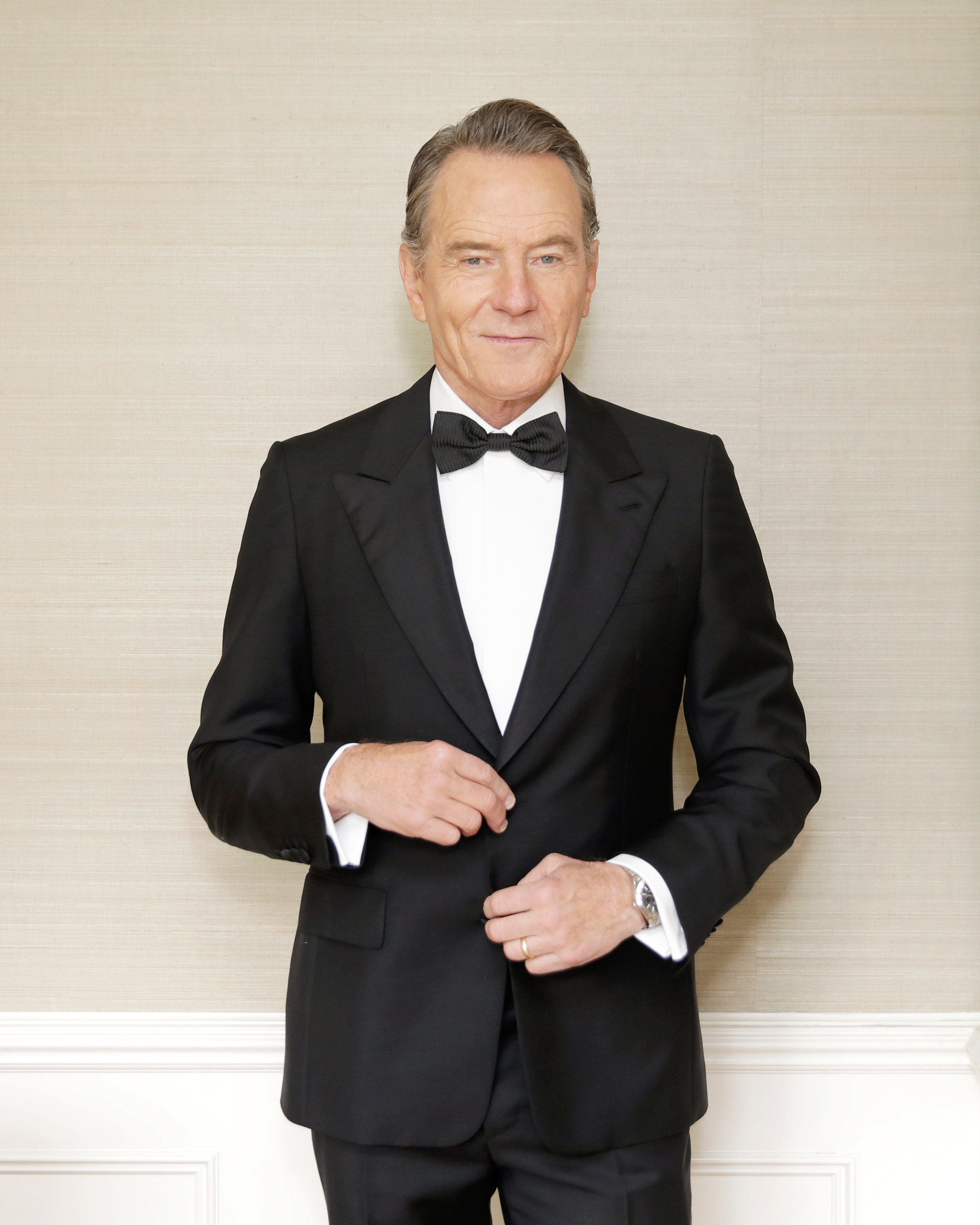 Bryan Cranston poses in front of a wall, wearing a buttoned-up tuxedo