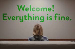 Eleanor Shellstrop sits in front of a painted sign that says, "Welcome! Everything is fine"