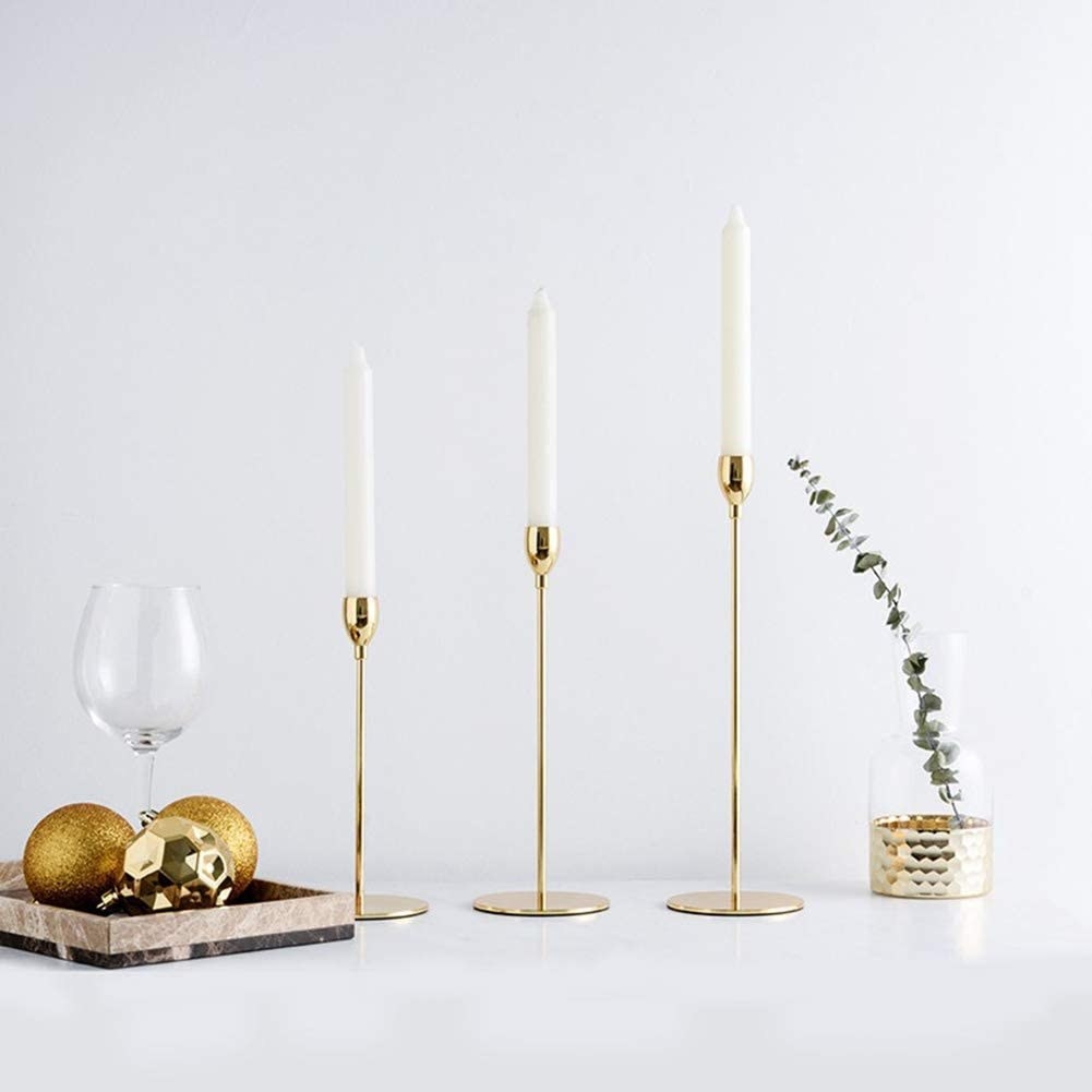 three taper candles in tall and slim candle stick holders on a white surface next to other home decor