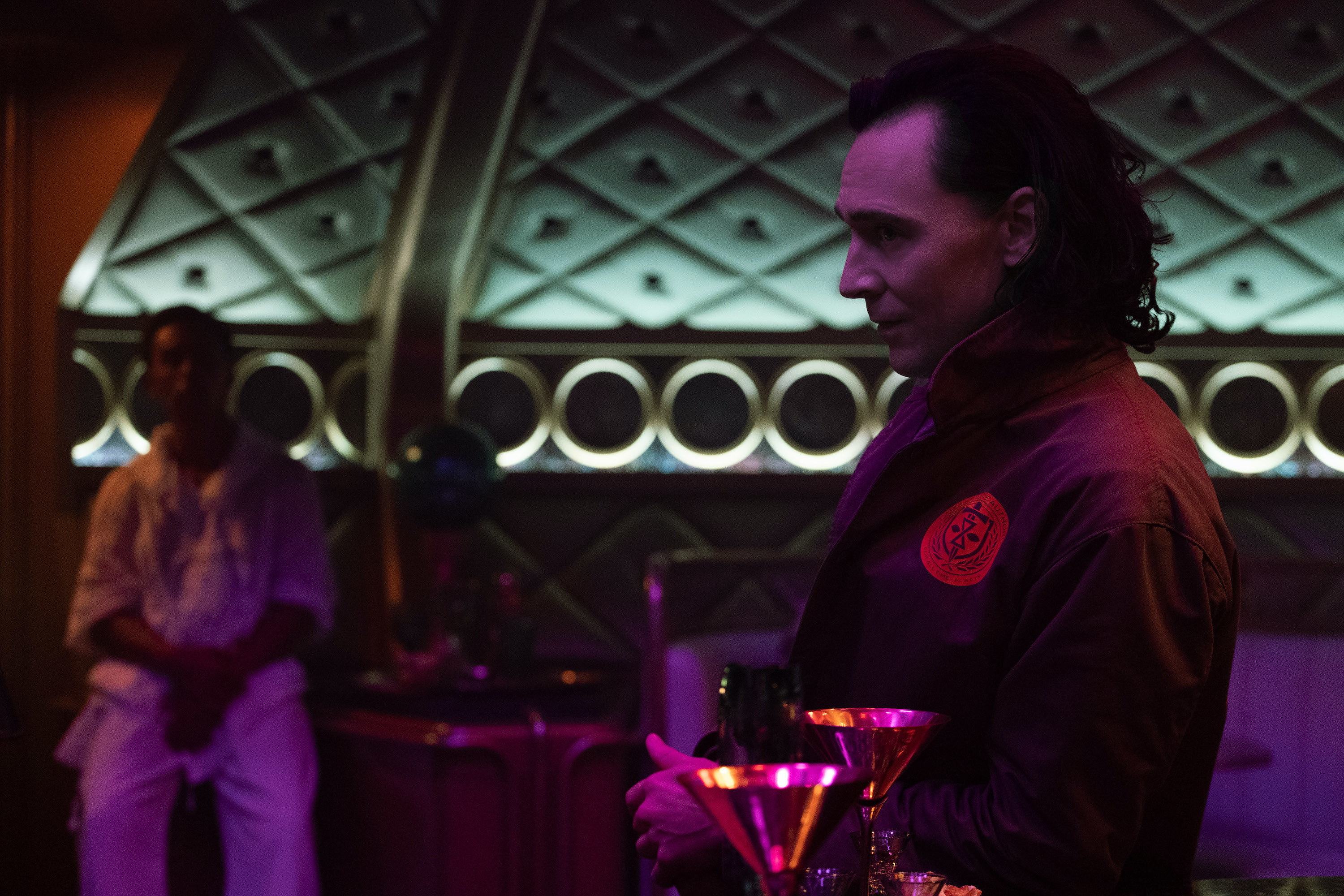 Loki slightly smirks while standing at a bar