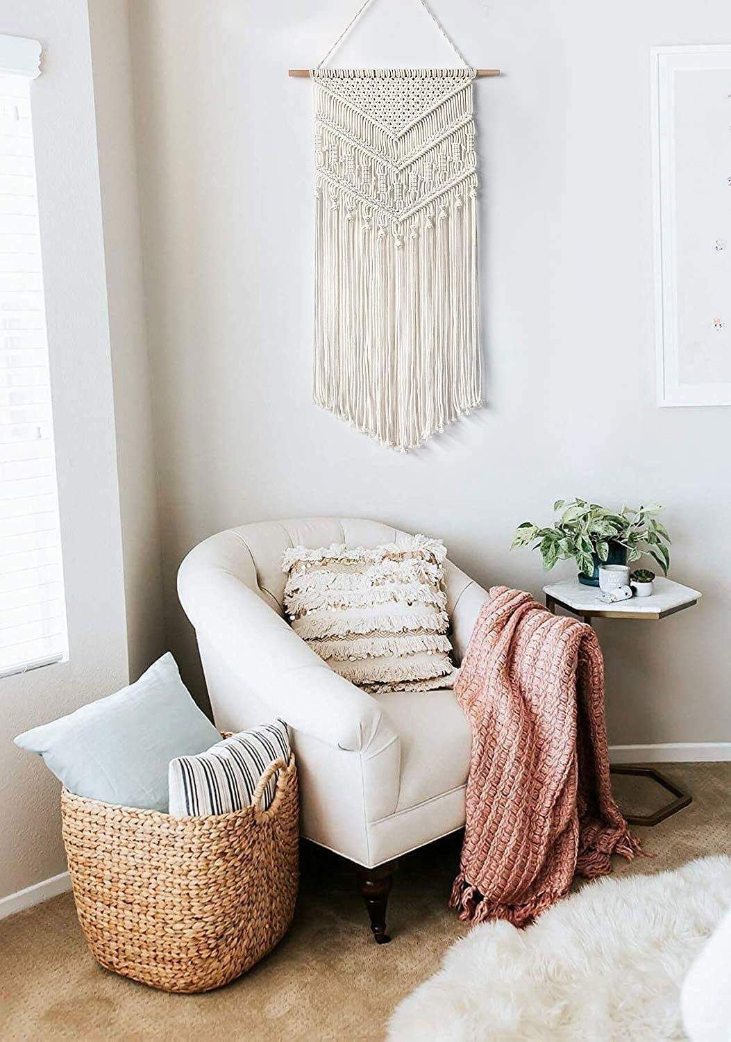 the macrame wall hanging on a wall behind a comfy chair and a basket of throw pillows
