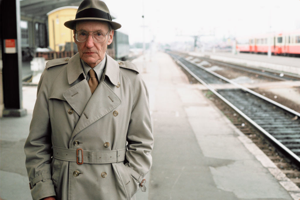 Burroughs stands at a train station in a trench coat and hat