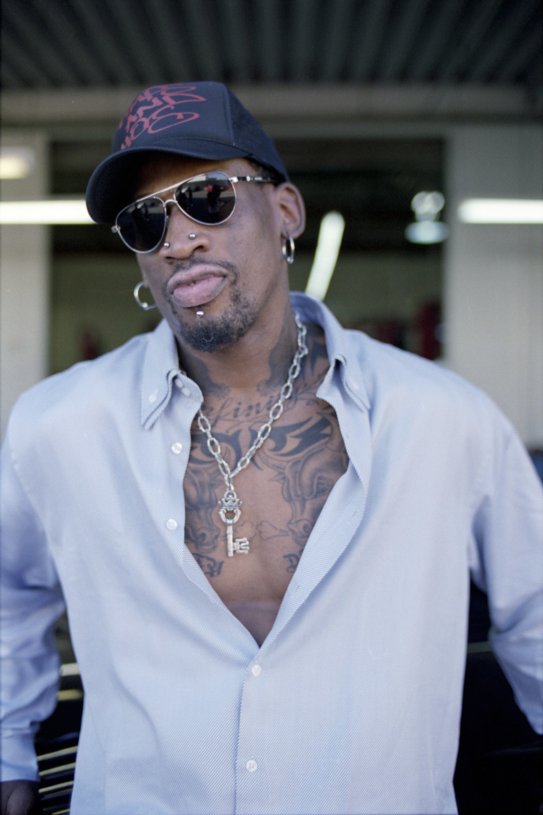 Dennis Rodman wears a hat and an unbuttoned blue shirt that shows off his chest tattoos