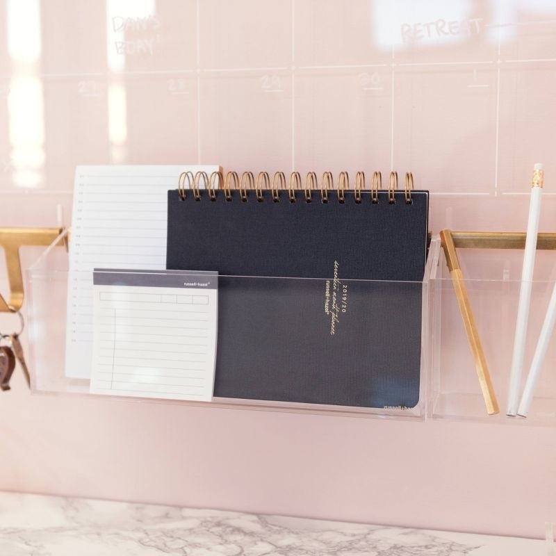 Clear wall organizer on pink wall holding notebooks and pens