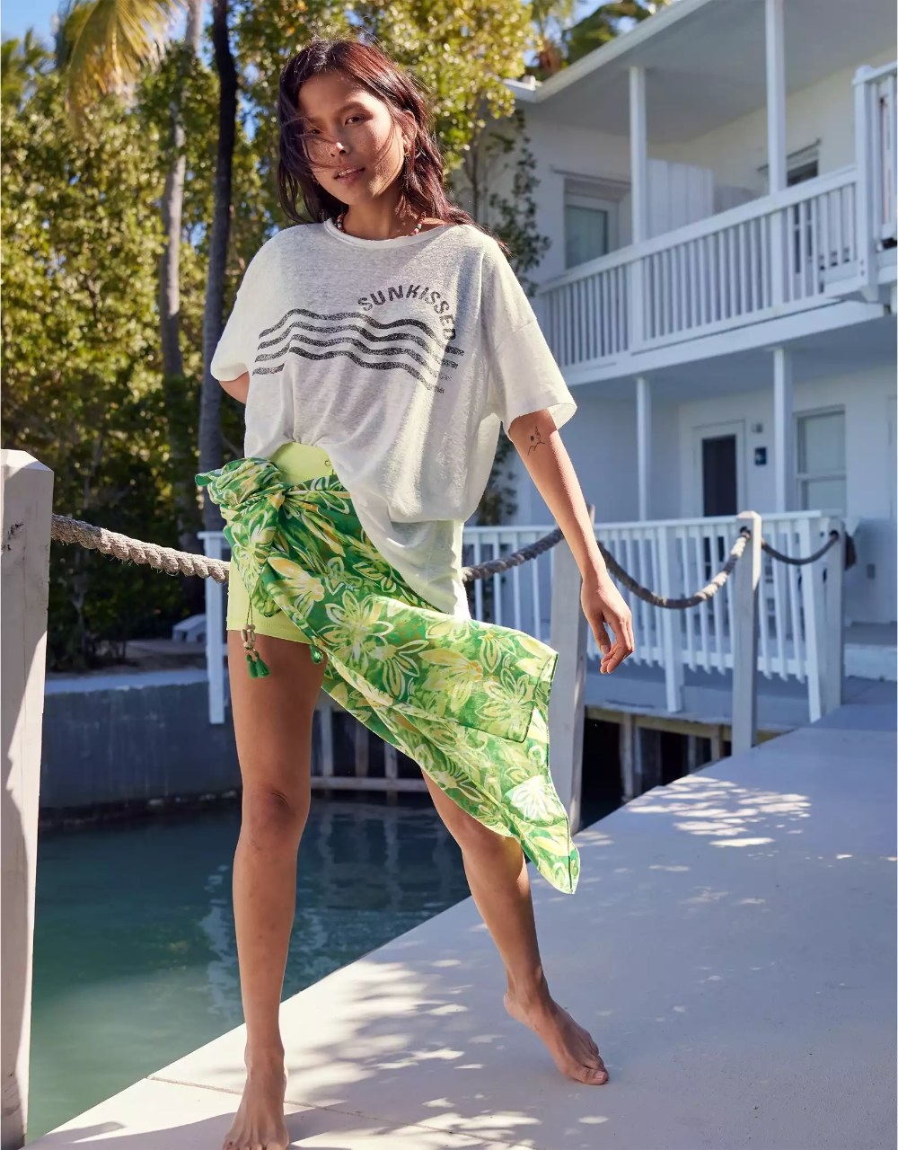 A model wearing the graphic tee in the color White, tucked into a green leaf print sarong