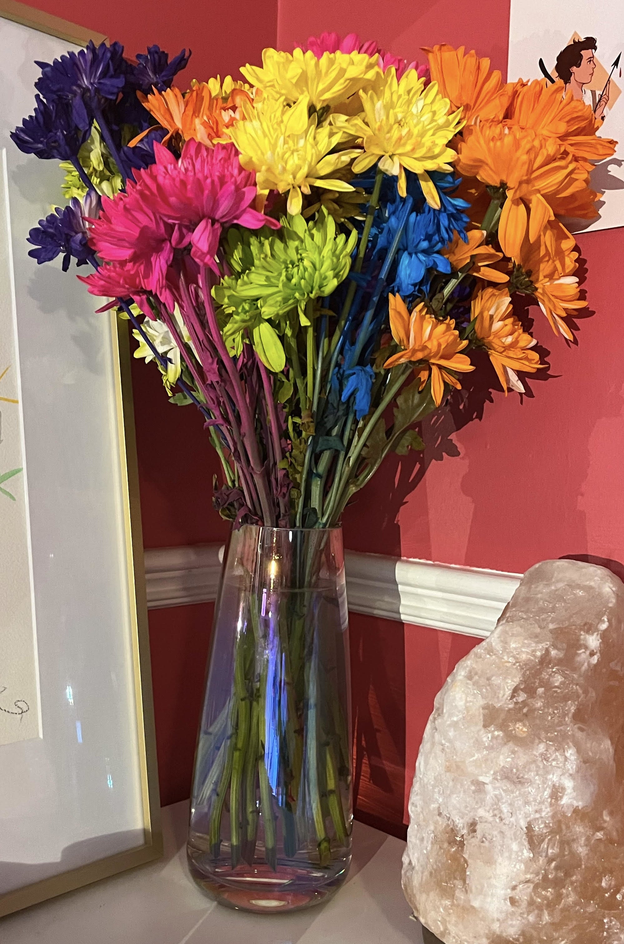 The vase on a table with flowers inside of it next to a painting and a salt lamp