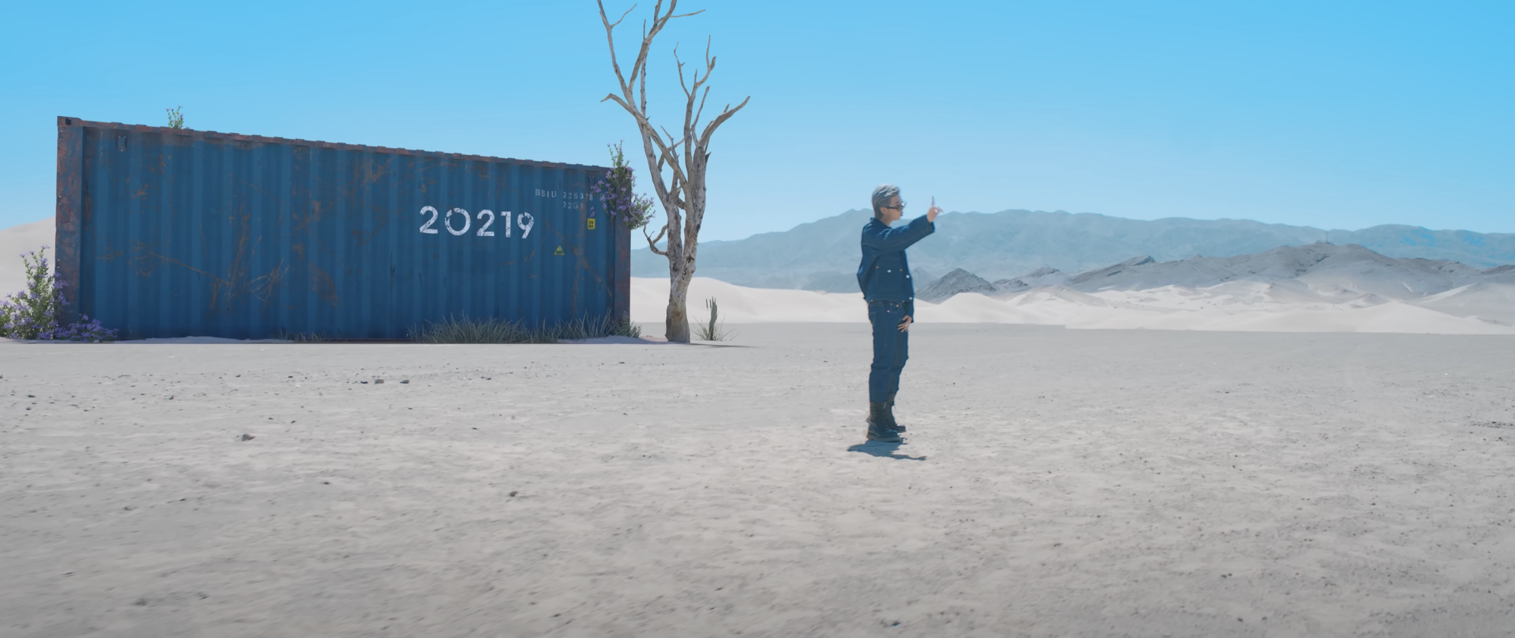 A still from a music video shows a cargo container with the number 20219, a dead tree, and a person with their hand up in a white sandy desert