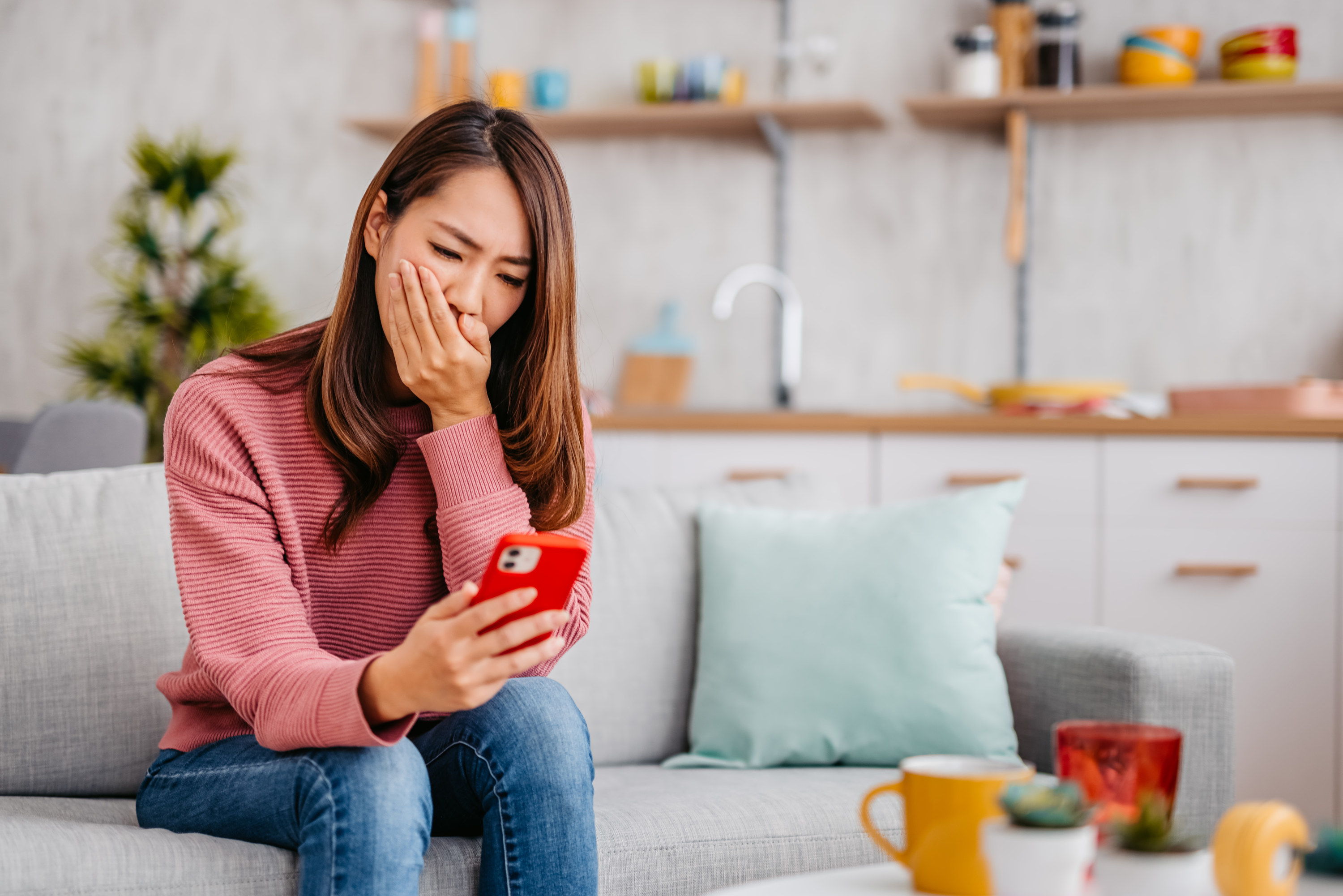 Woman sitting on a couch and looking at her phone while covering her mouth with her hand
