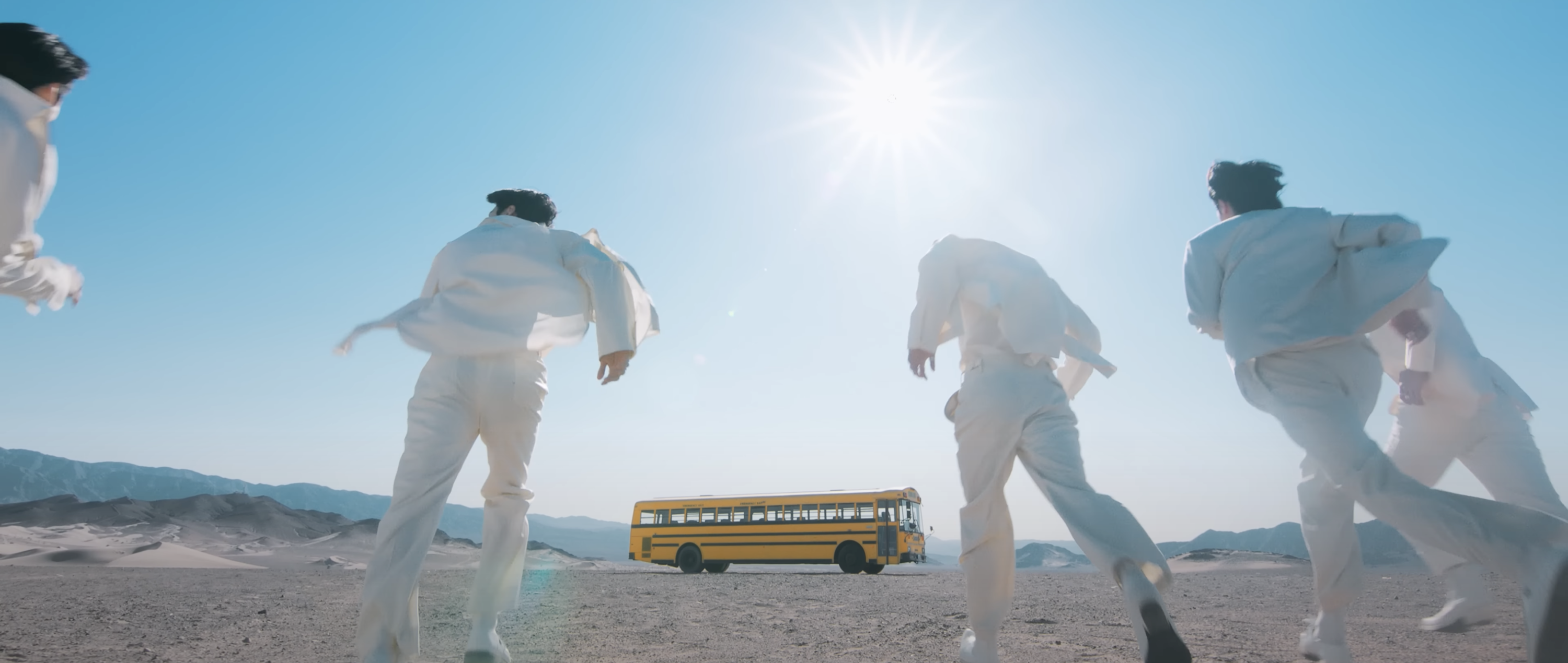 A still from a music video shows members of BTS dressed in white in a desert by a school bus in the distance