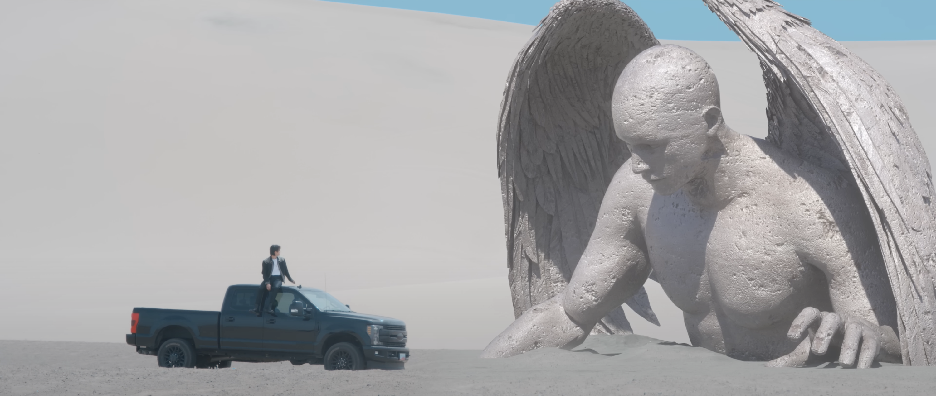 A still from a music video shows a member of BTA sitting on a truck facing an angel statue in the desert