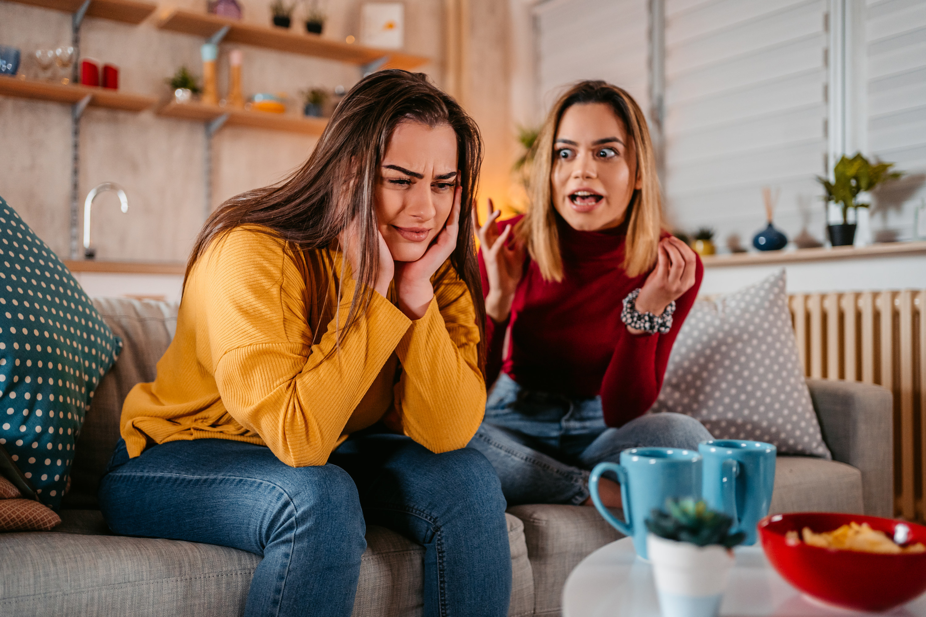 Two women sitting on a couch, one with her head in her hands and the other saying something to her