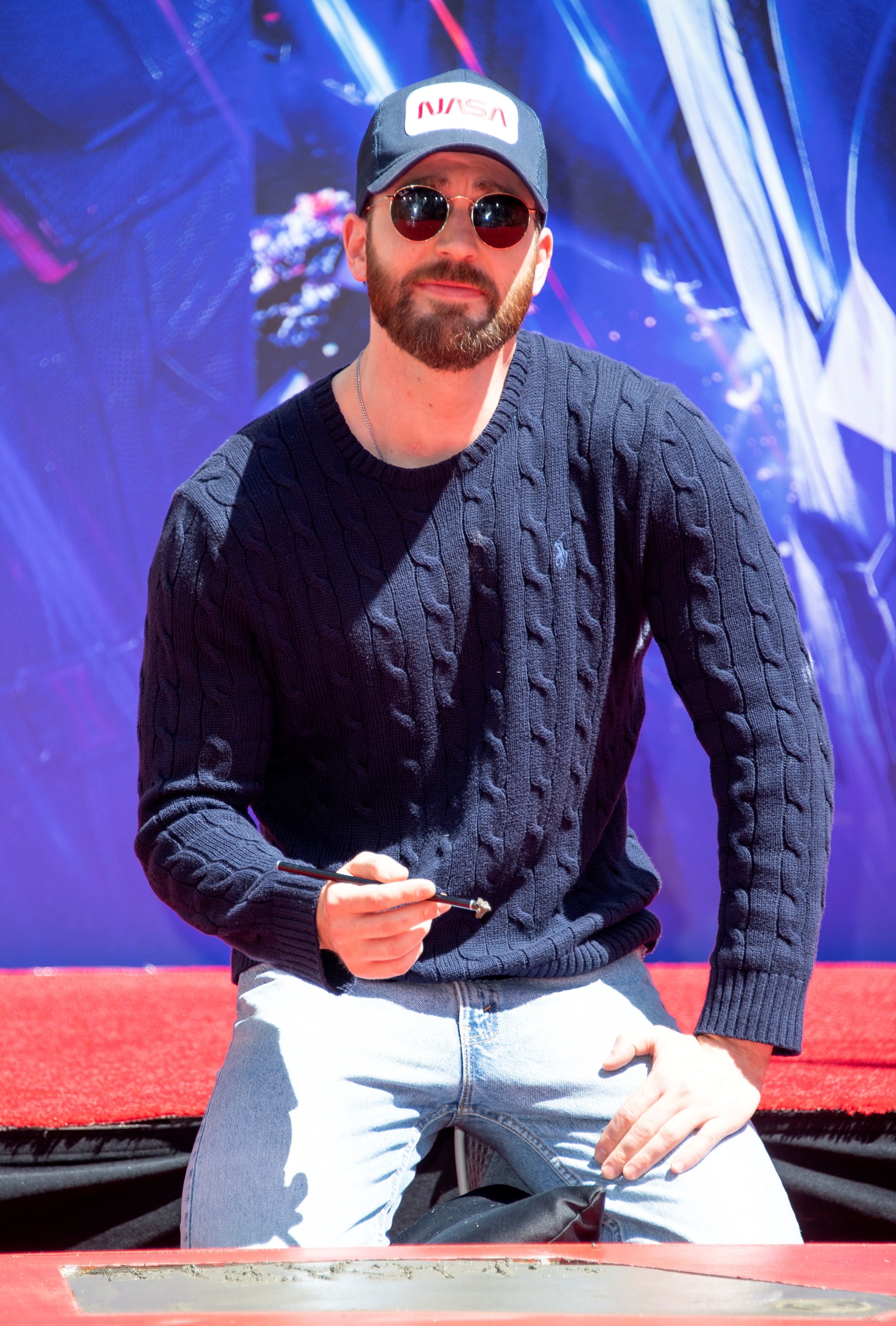 Chris on his knees in a cap, sunglasses, sweater, and jeans