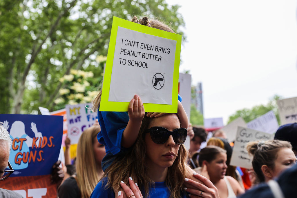 Child on a woman&#x27;s shoulder holds up the sign &quot;I can&#x27;t even bring peanut butter to school&quot; with a gun crossed out