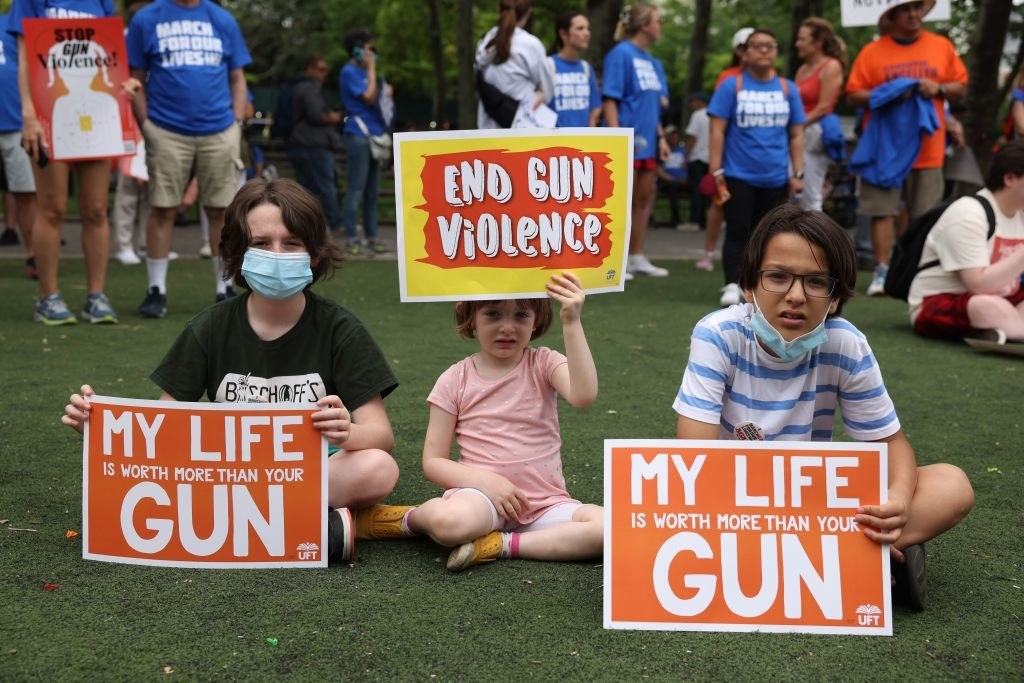 Three children sitting and holding signs, including &quot;End gun violence&quot; and &quot;My life is worth more than your gun&quot;