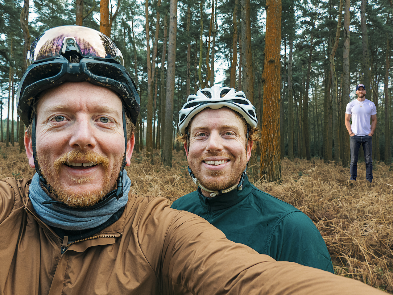 chris in the foreground of two cyclists taking a selfie in the woods