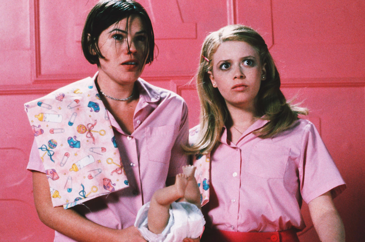 two women standing in a lurid pink room wearing pink outfits and looking wide eyed at something off camera
