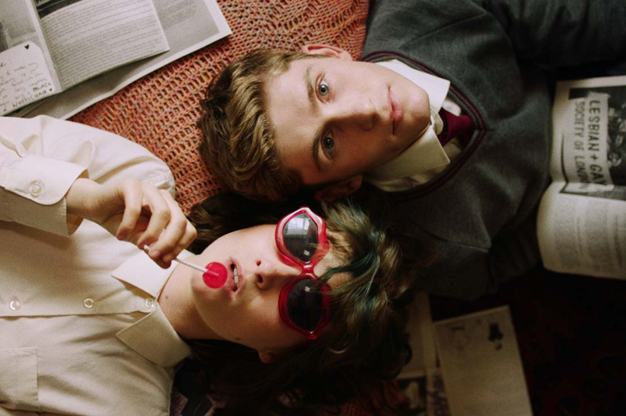 two teens lie down in opposite directions the girl wear sunnies and has a lollipop the guy looks worried and holds a book