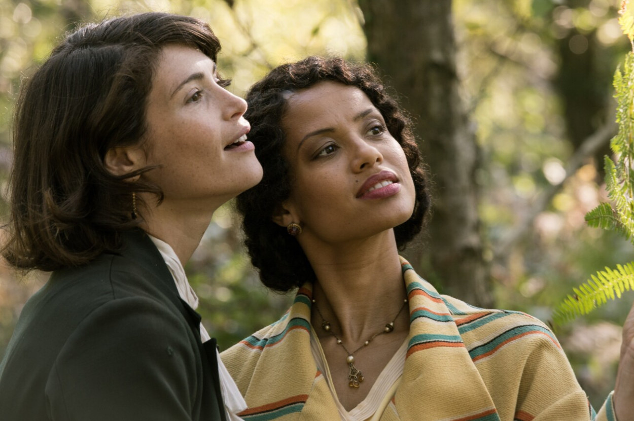 Two women stand in a forest area and gaze as something off camera they look like they are holding each other