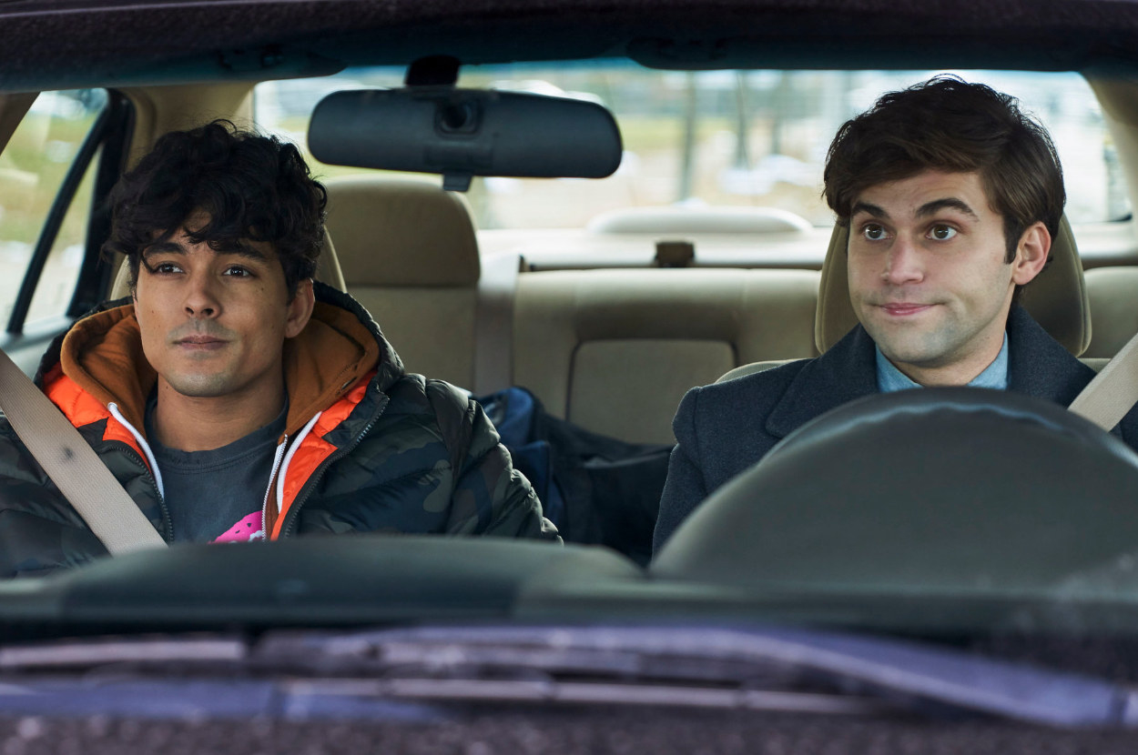 Two young men sit in a car together side by side looking bemused