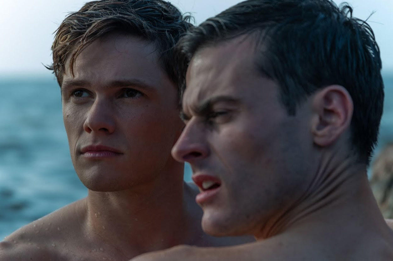 two men hold each other close near a rock in the ocean they are both topless and looking at something in the distance