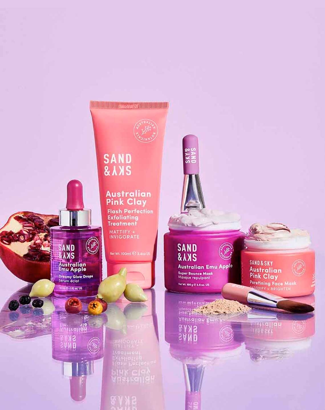A pomegranate and other yellow and pink fruits with a jar of purple liquid, a tube of pink products, a purple tub with an applicator and a pink tub