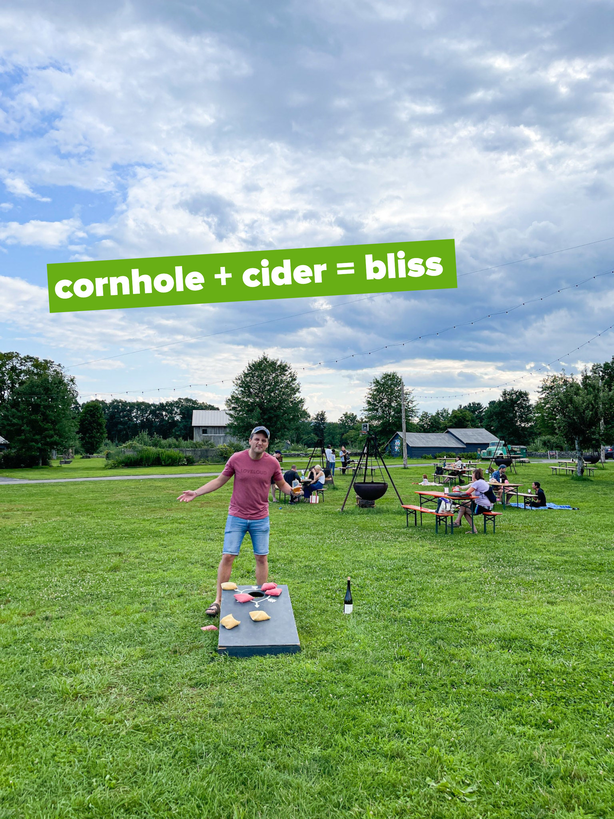 A man standing amid a field with people at outdoor tables and a fire pit with the text &quot;cornhole + cider = bliss&quot;