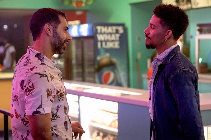 Two men look at each other affectionately near the kiosk of a movie theatre snack bar