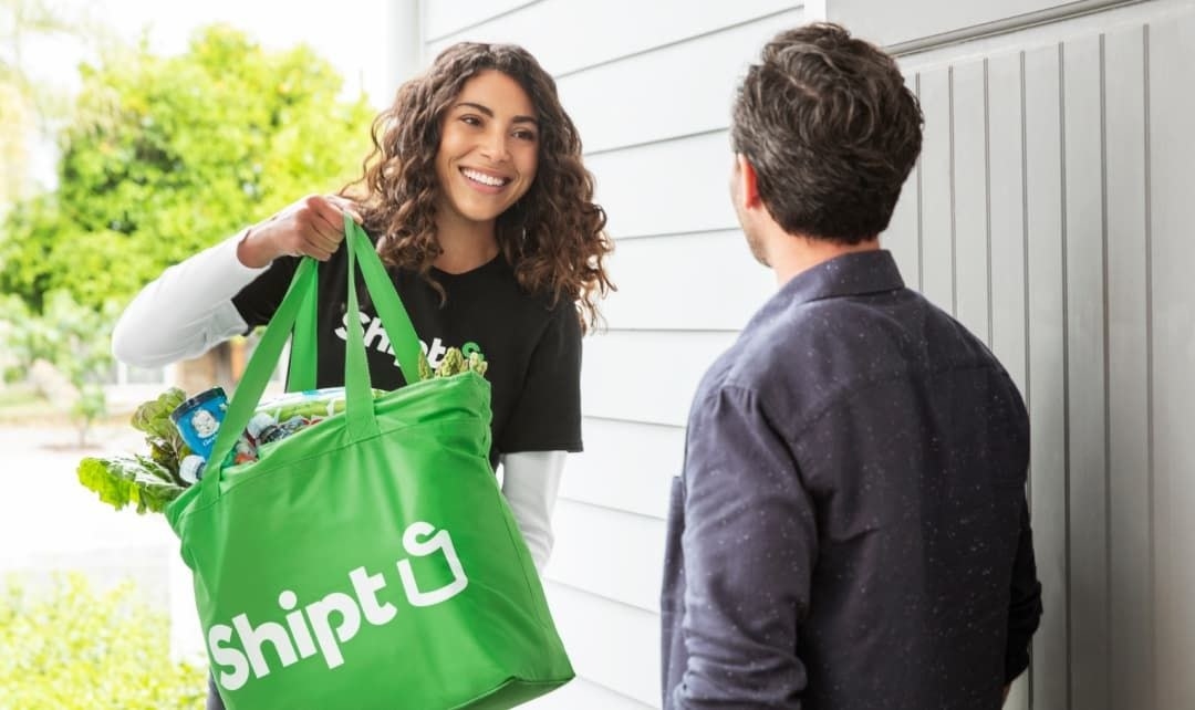 a person handing a green grocery bag that reads &quot;shipt&quot; to another person