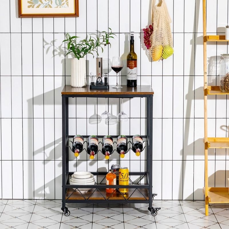 Black, rectangular bar cart against kitchen wall. It&#x27;s holding wine bottles, glasses, dishes, mixers, and a decorative plant.