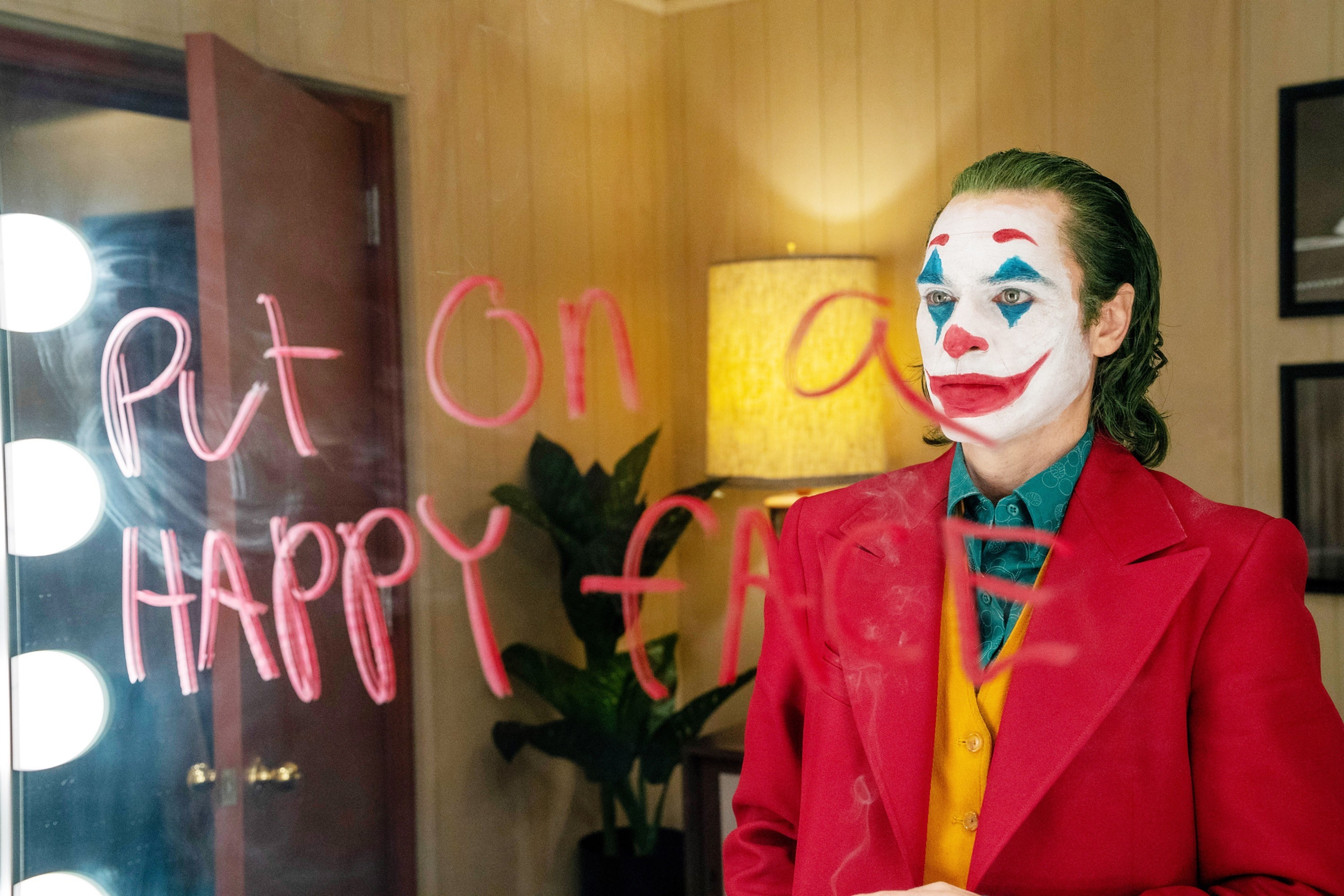 Joker from the first film standing in front of a mirror that says &quot;put on a happy face&quot;