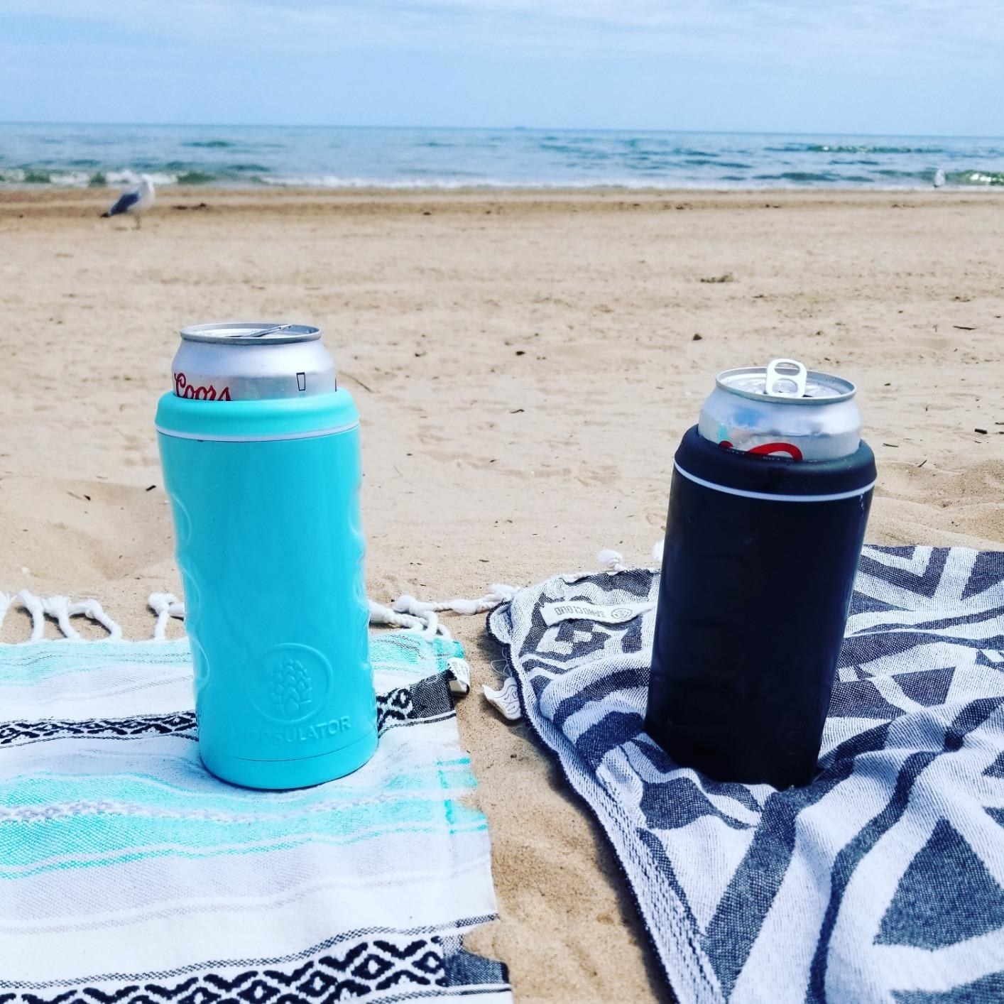 two cans (one blue, one black) holding sodas on the beach