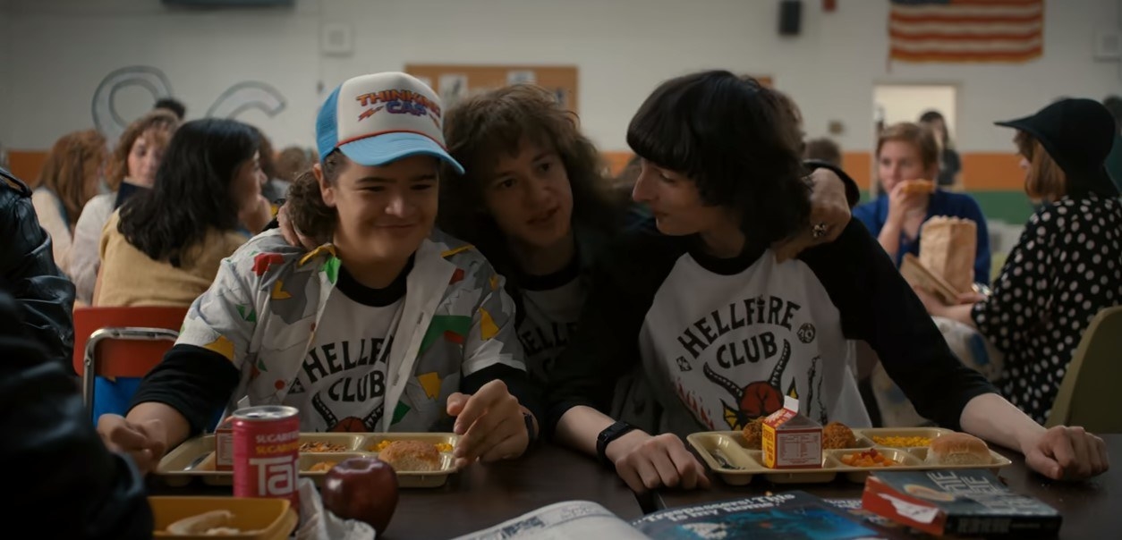 The image shows Dustin, Eddie and Mike wearing Hellfire Club t-shirts at a school canteen table