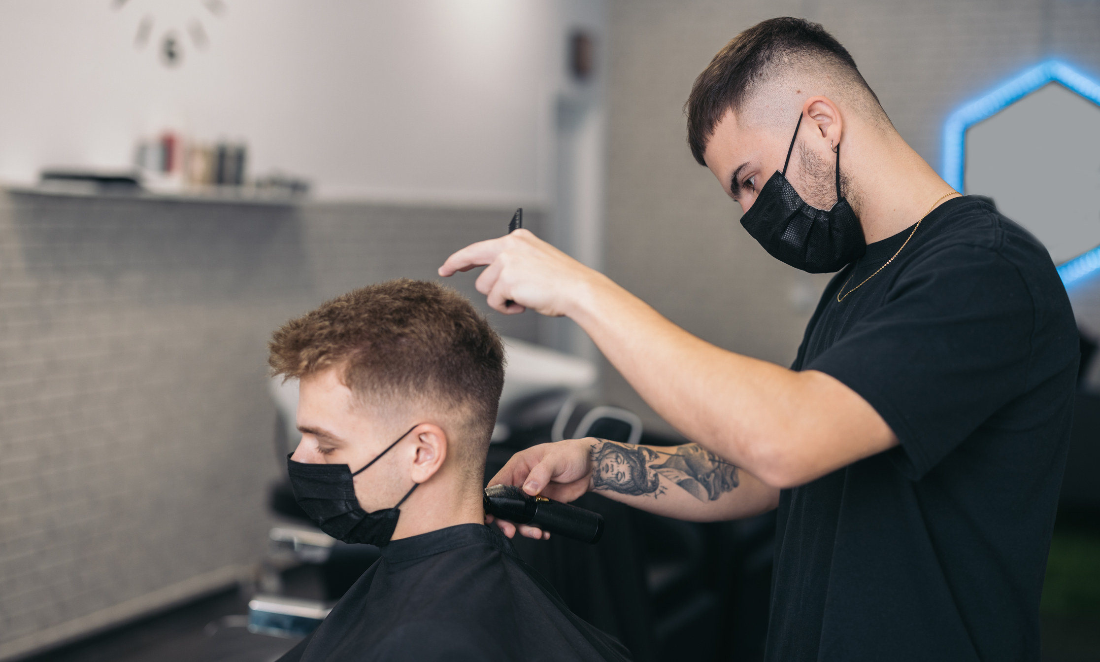 A barber wearing a surgical mask works on a young man who&#x27;s also wearing a mask