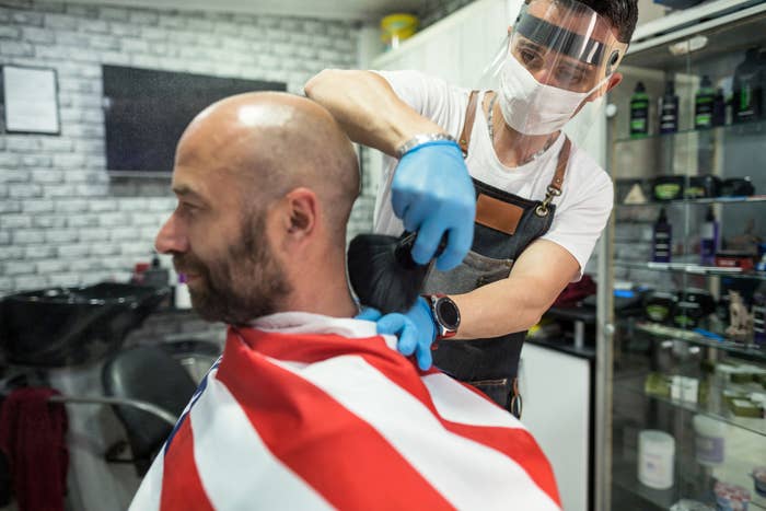 Barber wearing a surgical mask, gloves, and a face shield brushes hair off a customer&#x27;s neck