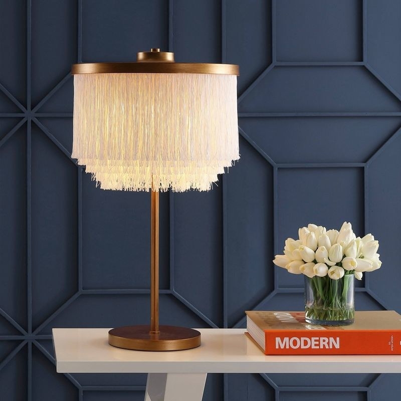 Lamp placed on white table next to a book and vase of flowers, against dark blue wall backdrop